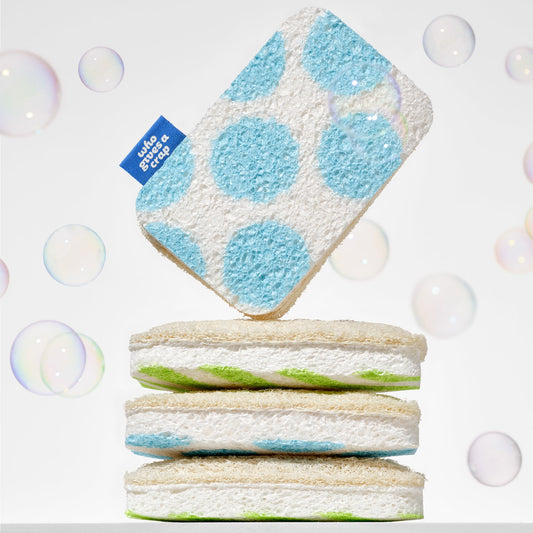 A Who Gives A Crap blue polka dot scrub sponge balanced on top of a stack of green striped sponges surrounded by soap bubbles