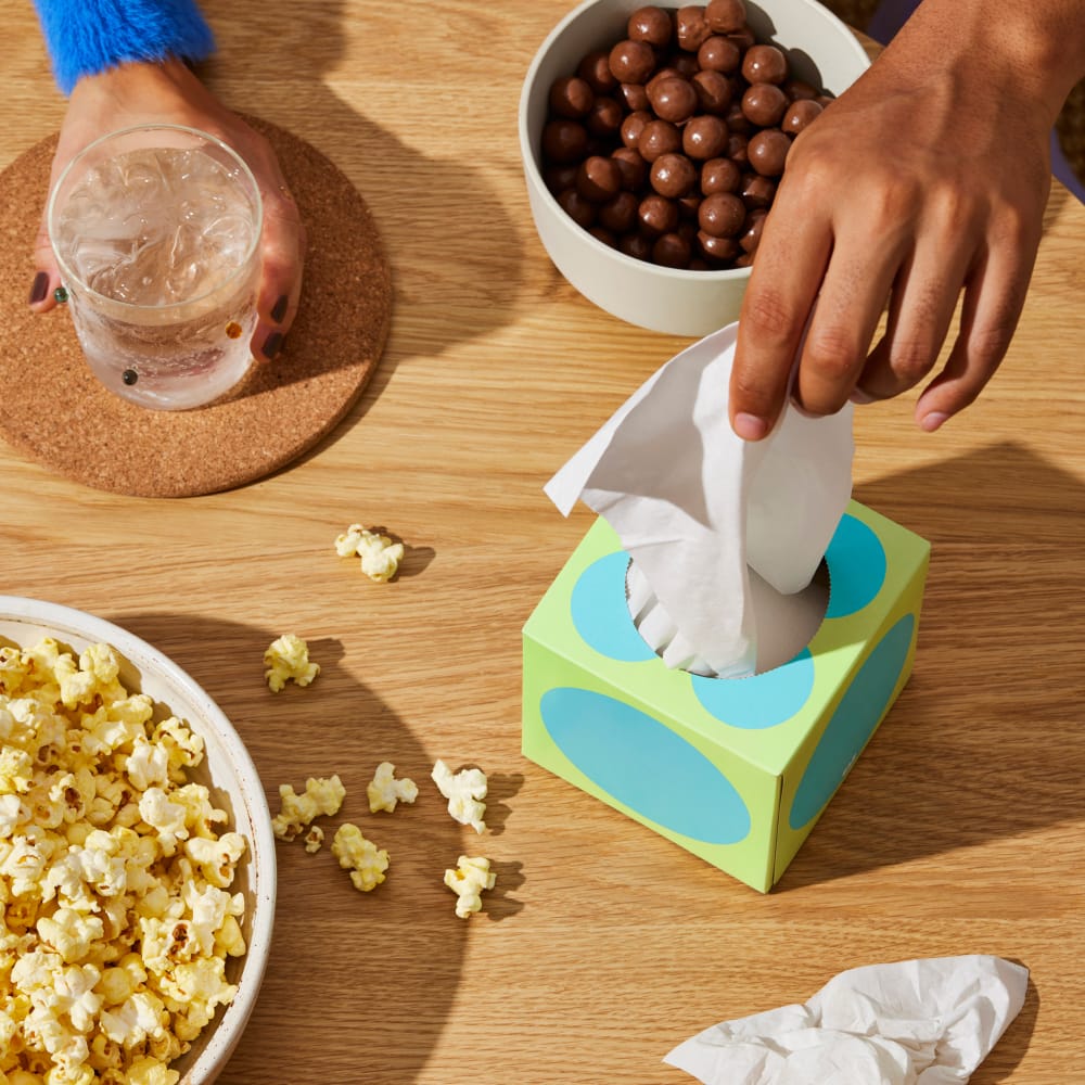 Hand grabs tissue from Who Gives a Crap tissue box on table with snacks and drinks