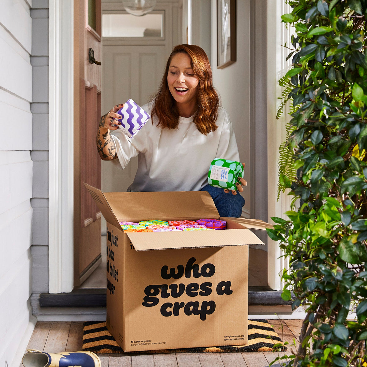 Woman collecting box of Who Gives a Crap toilet paper from front porch of home smiles as she opens box