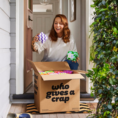 Woman collecting box of Who Gives a Crap toilet paper from front porch of home smiles as she opens box