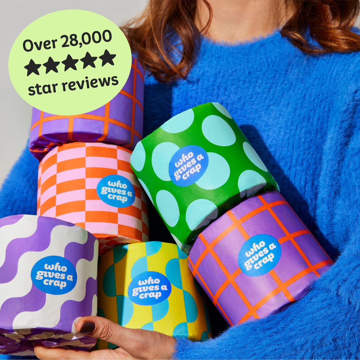 Woman holds handful of colourful Who Gives a Crap recycled toilet paper rolls. Copy over the image reads ‘Over 28,000 5 star reviews’.